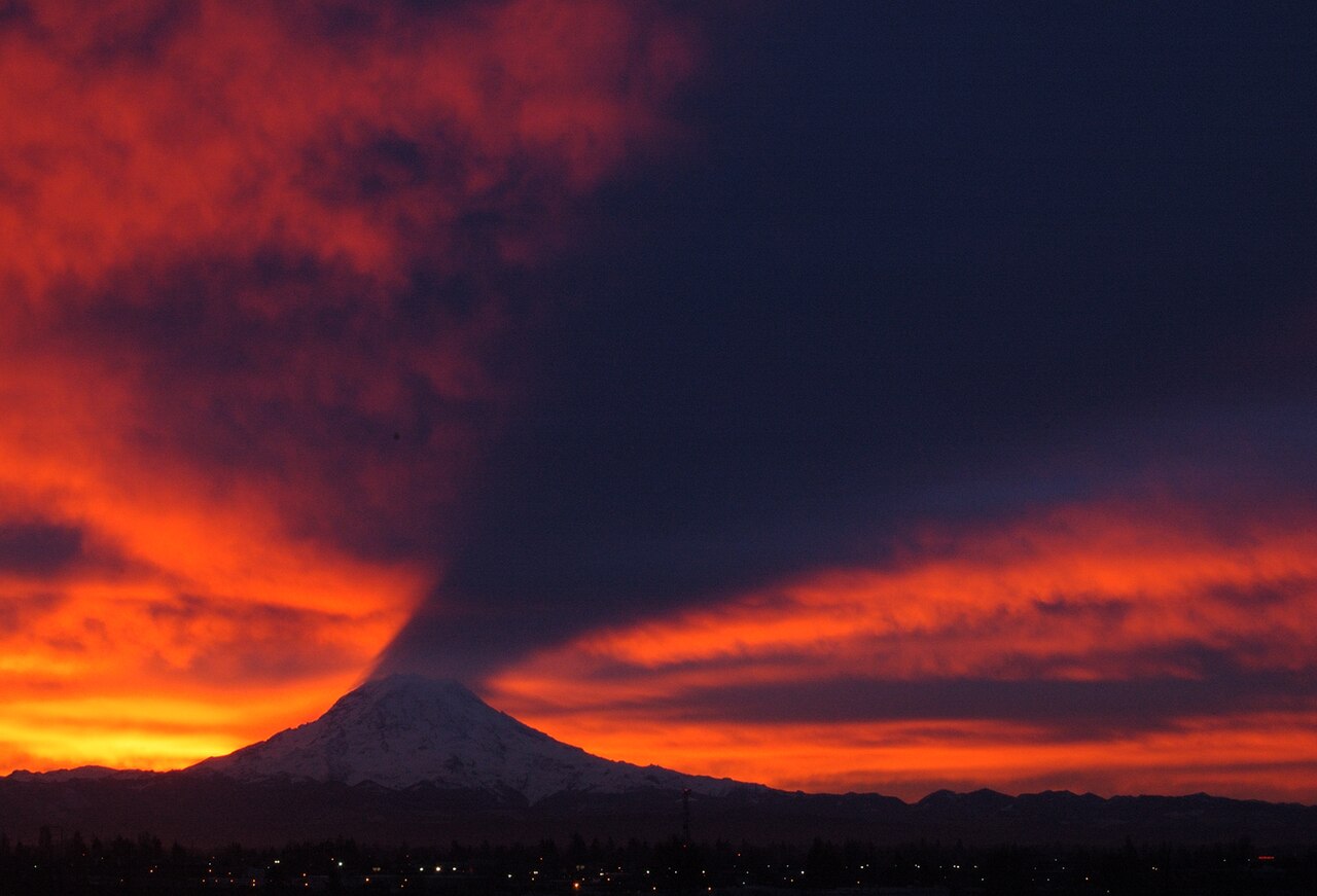 El monte Rainier al amanecer proyectando un cono de sombra oscuro hacia arriba sobre la capa de nubes, contra un cielo naranja