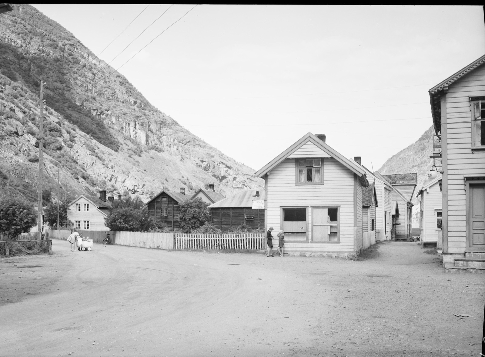 Una calle de tierra en Lærdalsøyri flanqueada por casas de madera blanca, con unos niños y un cochecito al borde del camino y la pared de roca desnuda del fiordo alzándose justo detrás de los tejados