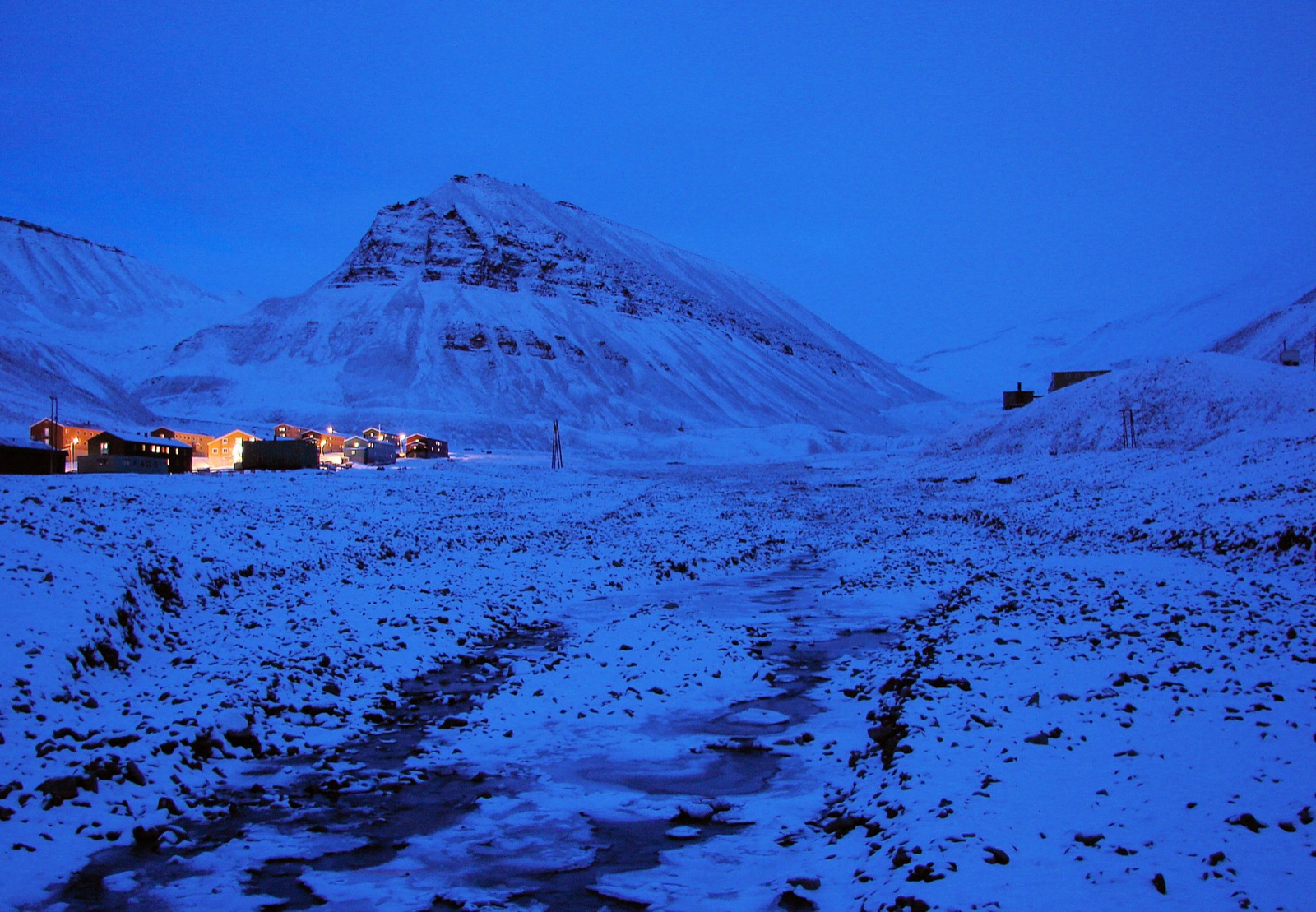 Longyearbyen en penumbra polar: una hilera de casas con ventanas iluminadas sobre la nieve, bajo una montaña piramidal oscura, todo bañado en un azul profundo