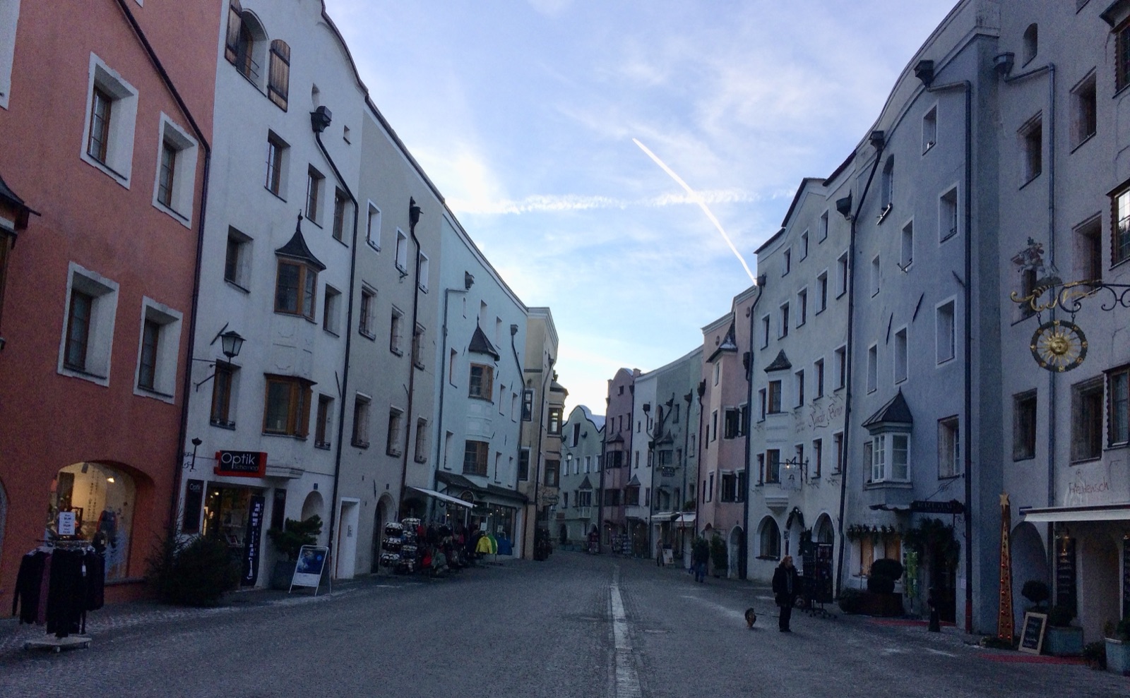 La calle principal de Rattenberg dos días después del solsticio de invierno: casas altas y pintadas a ambos lados, luces encendidas en las tiendas, una mujer paseando un perro y todo el suelo de la calle en sombra bajo un cielo azul brillante
